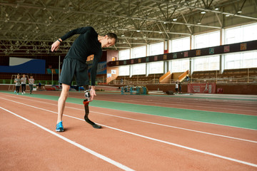 Full length portrait of young handicapped athlete with artificial foot warming up before running practice in modern gym stretching to sides, copy space