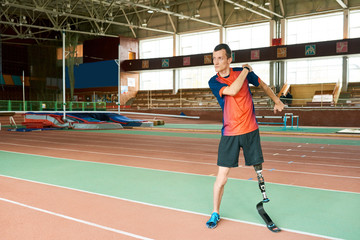 Full length portrait of young amputee sportsman warming up before running practice in modern indoor stadium , copy space