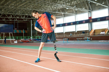 Full length portrait of young handicapped sportsman with artificial foot warming up before running practice in modern gym stretching to sides, copy space