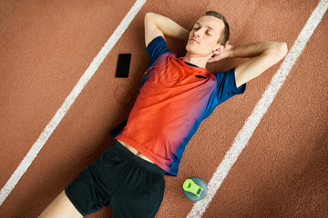 Above view portrait of young sportsman lying on running track with eyes closed, resting after practice and listening to music