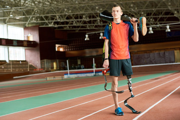 Full length portrait of young amputee sportsman standing in modern indoor stadium holding spare prosthetic leg, copy space