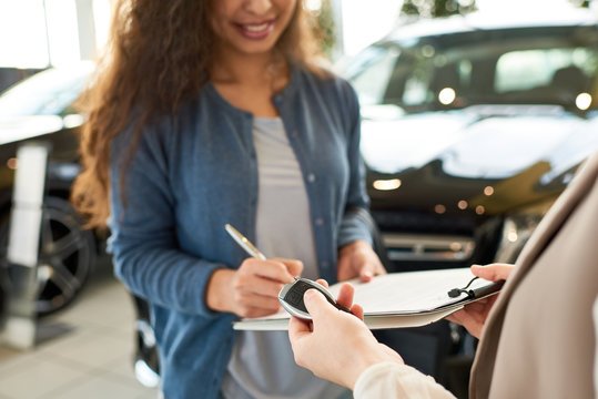 Close-up shot of smiling curly woman signing contract for new car, unrecognizable saleswoman holding key in hand, interior of modern showroom on background