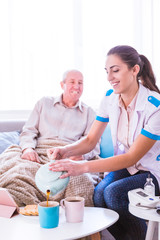 The young smiling doctor sitting near the old, ill, smiling man at home and pouring tea into the cups'