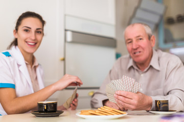 Portrait of the old joyful man and his young cheerful doctor who sitting at the table in the kitchen and they playing cards