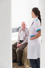 The old smiling man sitting on a bed in the room at home and the young nurse standing in a room near the door