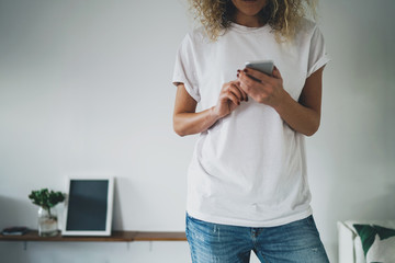 Fototapeta premium Cropped shot hipster girl wearing a white blank empty t-shirt and blue jeans using a mobile smartphone. Young woman writing text message on her cell phone. white wall background. Horizontal mockup.