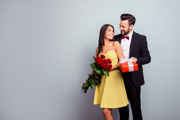 Portrait with copy space of attractive couple in formal wear, woman in dress holding bouquet of red roses, man in tux embracing her from back side and presenting a packaging red case with gift