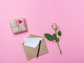 Pink rose, craft envelope with blank paper, pen and gift with pink heart on pink background. Love...