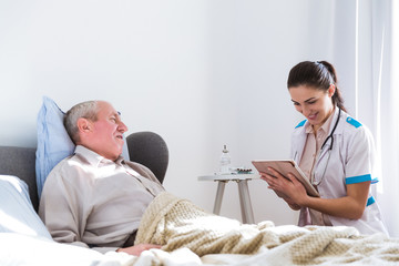Fototapeta premium The old, sick, man lying on a bed in the room looking at the young doctor who is sitting near him and looking at the tablet