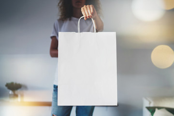 Young hipster girl wearing blank white t-shirt and holding white paper package with empty space for your logo or design, mock-up of shopping bag with handles. Bokeh light.