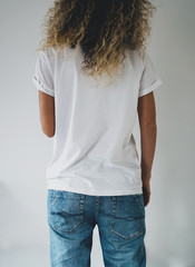 An attractive young woman dressed in a white blank t-shirt and blue jeans posing on a background of a white wall. Vertical mock up. empty space for you logo or design. Back view.