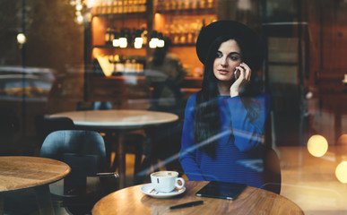 attractive girl with long black hair talking on the phone sitting in a restaurant in front of a large panoramic window. young business woman is waiting for a business partner for a business meeting