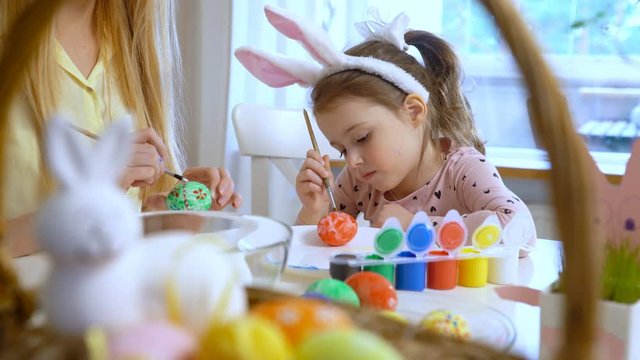 Happy Easter! Young mother and her cute little daughter wearing funny rabbit ears are coloring easter eggs sitting at a festive table with basket and Bunny