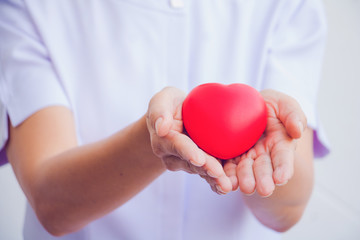 A nurse holding red heart toy. She is Left / right hand holding it. She is smile and good mood. The photo shows the principle of caring and good health.