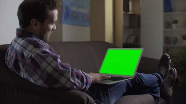 Man sitting on couch and using laptop with green screen, socializing on website