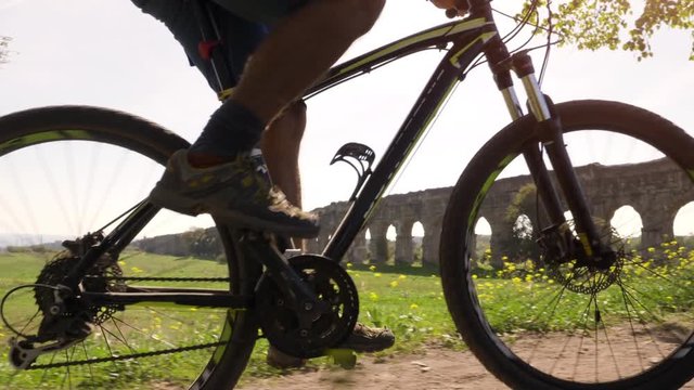 Cyclist riding bike in front of ancient roman aqueduct in a park. Young attractive athletic man with orange sportswear and backpack in parco degli acquedotti in Rome
