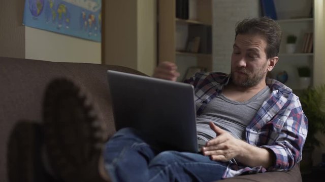 Untidy male lying on couch, looking at laptop laughing at what he sees, laziness
