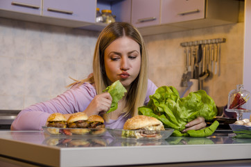 Woman eating salad and looking at tasty burgers on kitchen counter, dieting concept 