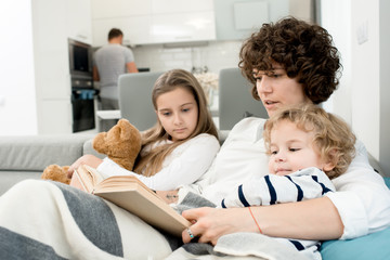Obraz premium Attractive curly woman sitting on cozy sofa with her adorable little son and daughter and reading them aloud while her husband preparing breakfast for all family.