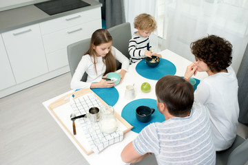 Naklejka na ściany i meble High angle view of loving family of four wearing pajamas sitting at kitchen table and enjoying appetizing breakfast