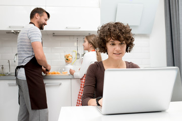 Obraz na płótnie Canvas Portrait shot of attractive middle-aged woman sitting at kitchen table and surfing Internet on laptop while her little daughter and bearded husband preparing healthy breakfast.