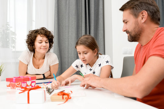 Preparing birthday present for granny: pretty little girl sitting at table with her middle-aged parents and drawing colorful picture with parents, gift boxes on foreground