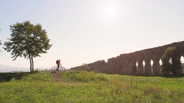 Cyclist with bike reads map for direction in front of ancient roman aqueduct in a park. Young attractive athletic man with orange sportswear and backpack in parco degli acquedotti in Rome under a tree