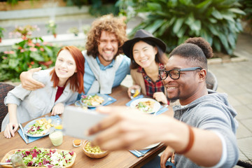 African-american guy with smartphone making selfie of him and his friends during festive dinner in the garden-room