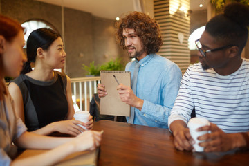 Handsome young businessman with notepad showing and explaining to colleagues what he thinks about new working points