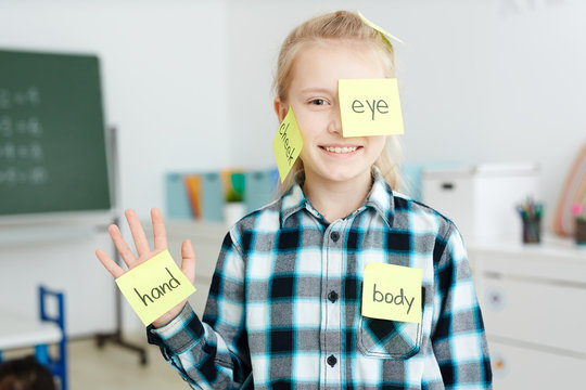 Happy schoolgirl having sticky notepapers with English names of body parts on her hand, cheek and eye