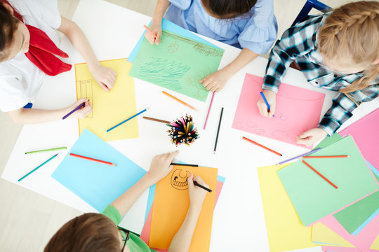 Overview Of Group Of Schoolchildren Drawing With Crayons While Sitting By Desk