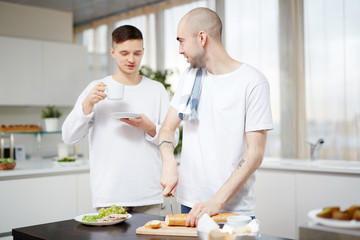 Young man cutting bread for sandwiches and talking to his partner in the kitchen
