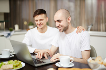 Contemporary homosexual couple sitting by table in front of laptop and watching online news