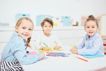 Happy little girls and boy looking at camera at drawing lesson in kindergarten