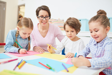 Happy teacher and three kids sitting by table at drawing lesson in kindergarten