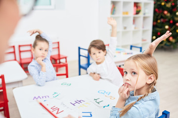 Three little children with raised hands sitting by table and looking to their teacher at lesson in...