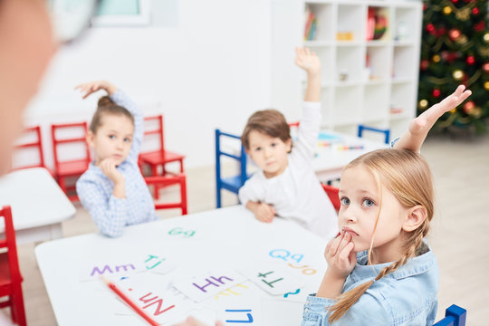 Three little children with raised hands sitting by table and looking to their teacher at lesson in kindergarten