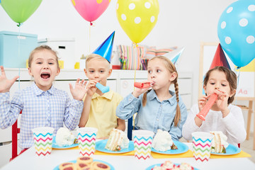 Ecstatic little friends with whistles sitting by birthday table and enjoying party