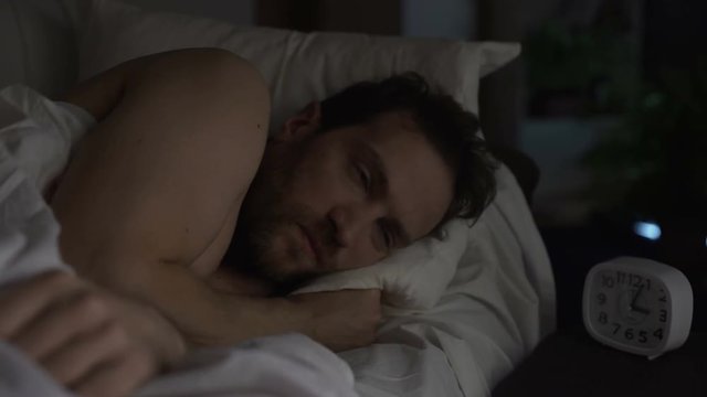 Guy turning in bed unable to fall asleep, looking angrily at clock, insomnia