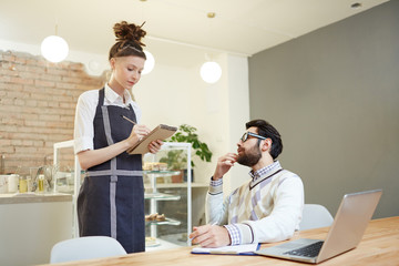 Young waitress with notepad and pencil writing down order of client in cafe