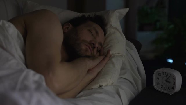 Bearded man sleeping on sofa bed, clock standing on night table, late night rest