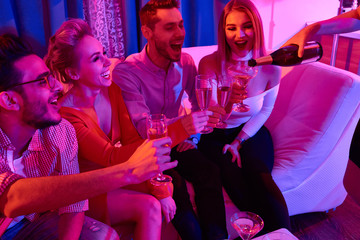 Group of glamorous  people enjoying private party, sitting on sofa laughing loudly while waitress pouring drinks