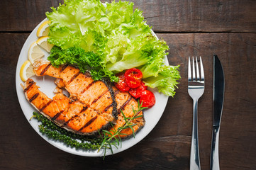 Grilled salmon steak served with grilled tomatoes, salad, lemon sliced, pepper seeds, rosemary, parsley on white plate on wooden table.