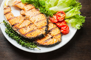Grilled salmon steak served with grilled tomatoes, salad, lemon sliced, pepper seeds, rosemary, parsley on white plate on wooden table.