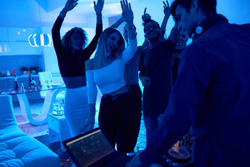 Group of modern young people dancing listening to DJ playing music at private house party, lit by...