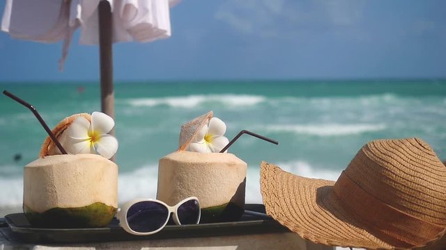 Tropical fresh coconut cocktails decorated frangipani flowers and sunglasses with hat in beach cafe with turquoise sea background. slow motion. 1920x1080