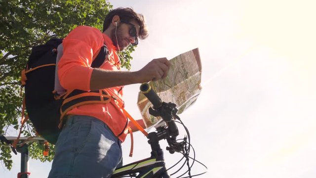 Cyclist with bike reads map for direction in front of ancient roman aqueduct in a park. Young attractive athletic man with orange sportswear and backpack in parco degli acquedotti in Rome under a tree