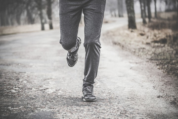 Young man running away on road outside of city in the dark cloudy afternoon. Daily outdoor active lifestyle. Enjoying sport concept. Runner's legs with sneakers.