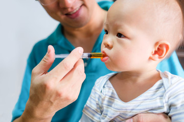 Cute little Asian 1 year old toddler baby boy child feeding with liquid medicine with a syringe, Dad giving remedy to kid who having flu, Fever cold and flu concepts. Selective focus at hand & syringe