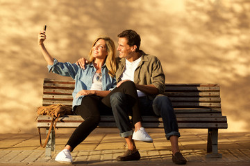 Full length happy couple sitting on park bench taking selfie